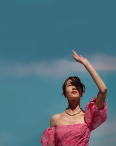 Liliya Chiang reaching up shot from below in pink dress in front of an almost clear blue sky