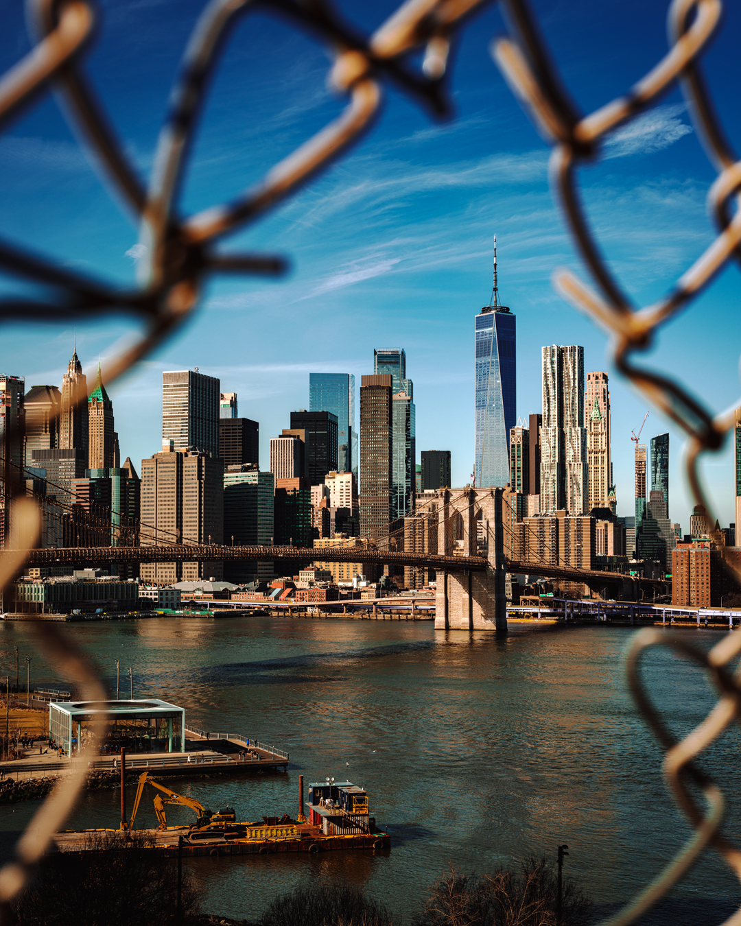 View of lower Manhattan framed by a break in the chain linked fence on the Manhattan Bridge